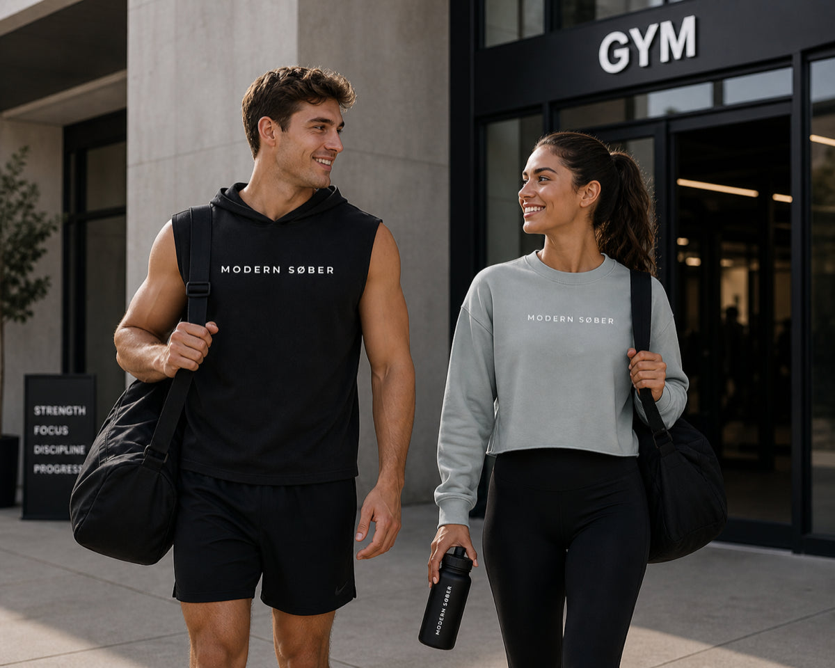A man and woman walking out of a gym smiling, both wearing Modern Søber apparel — black sleeveless hoodie and a sage cropped sweatshirt