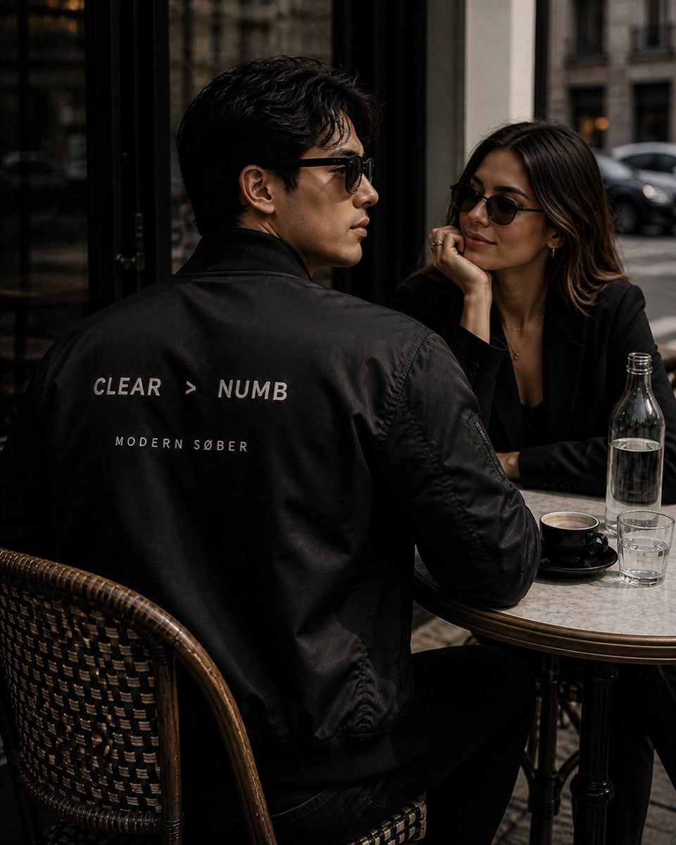 A couple at a cafe — man in Modern Søber CLEAR > NUMB black bomber jacket seated with back to camera, woman across from him resting her chin on her hand